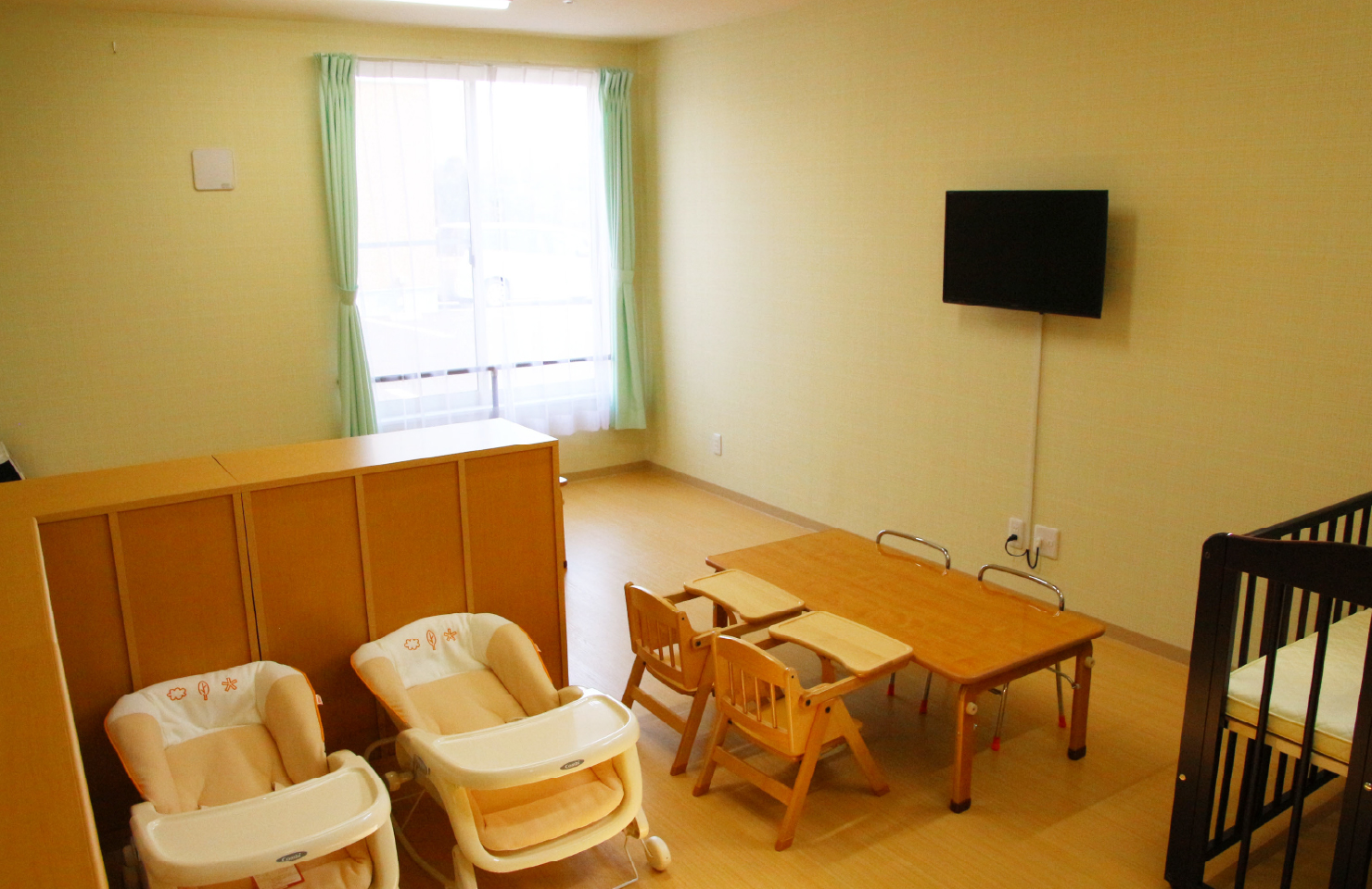 介護施設の保育スペース。ベビーベッドとテーブル、テレビが設置された明るい共用部屋。