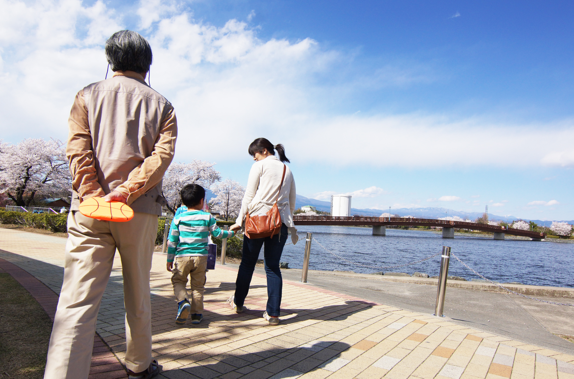 介護施設の入居者が河川沿いを散歩している光景。スタッフが付き添い、桜が咲く季節の屋外活動。